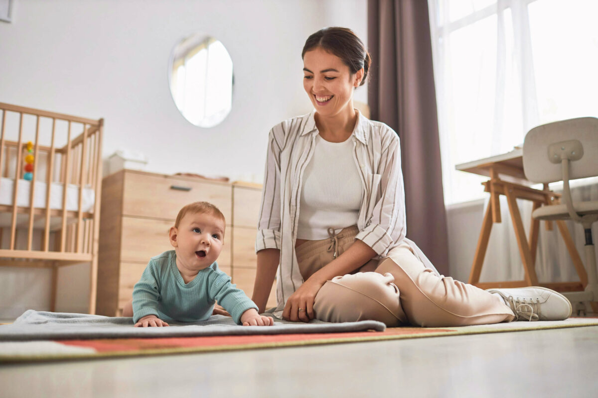 Ein Baby krabbelt auf einer Krabbeldecke, seine Mutter sitzt daneben und lächelt